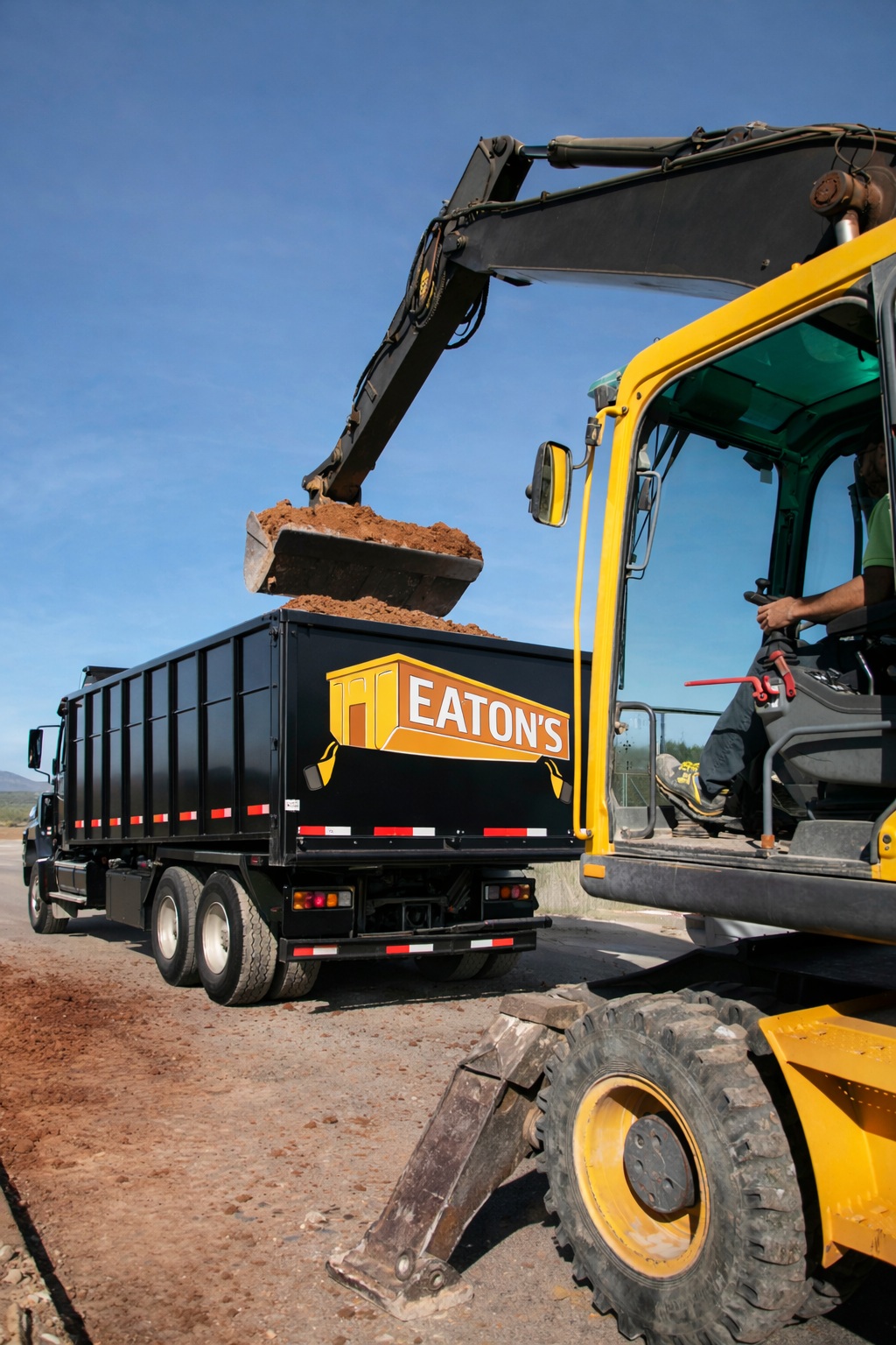 Excavator loading soil into Eaton’s dumpster truck at construction site, heavy-duty roll-off container for debris removal and earthmoving projects.