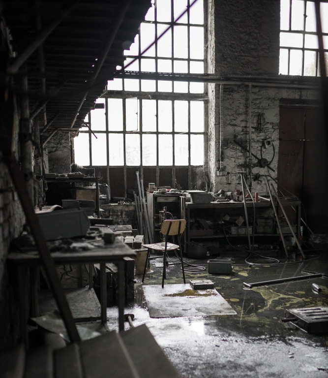 Abandoned industrial workshop interior with large windows, scattered tools, and damaged flooring in old factory building.